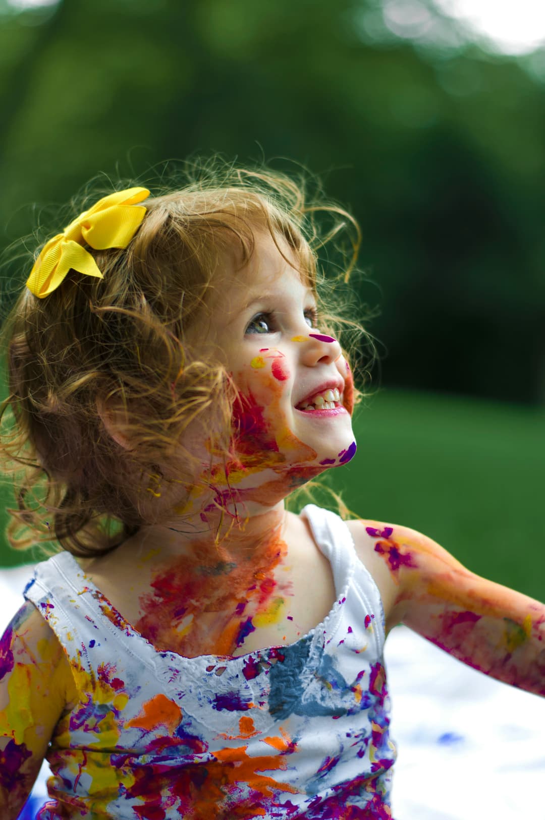 A little girl covered in colorful paint with a yellow bow on her head
